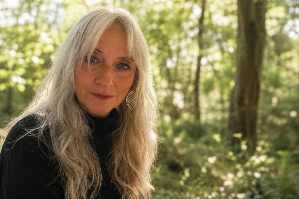 Portrait of Vanessa Champion, founder of Virtual Nature Walls, with long grey hair and leaf-shaped earrings, standing in a green woodland.