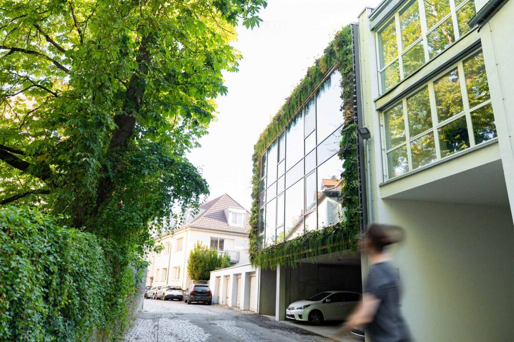 Voorbijganger bij een gebouw met een groene glazen gevel, links een kastanjeboom en een haag